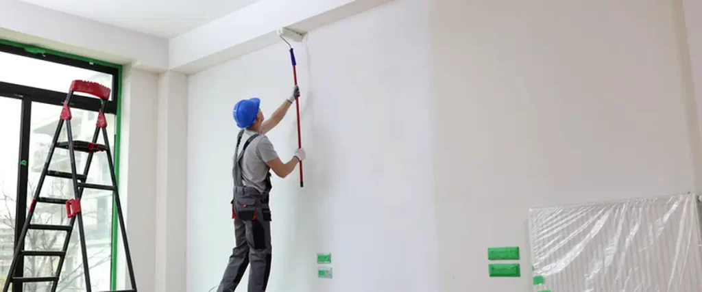 professional house painter in a blue hard hat expertly applies white paint to a large interior wall