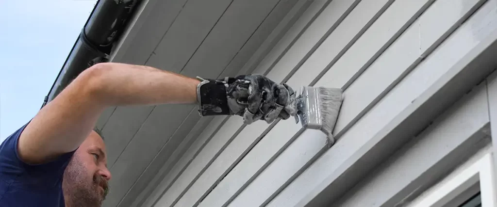 Painter using brush to apply light gray paint on exterior siding